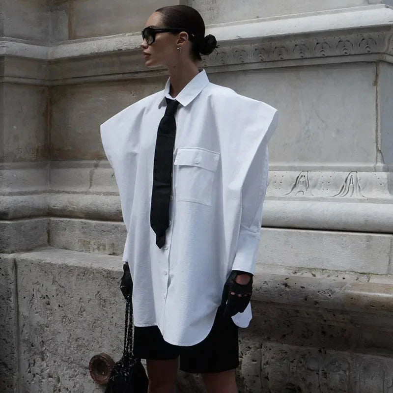 Person wearing a white oversized shirt with a black tie and shorts, standing against a stone wall.