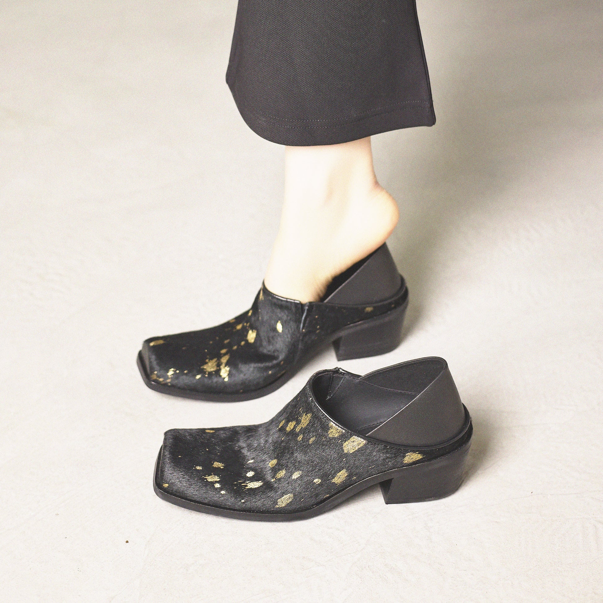 Black clogs with gold speckles worn by a person on a neutral background