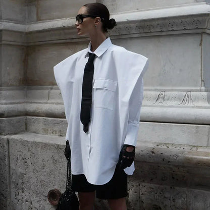 Person wearing a white oversized shirt with a black tie and shorts, standing against a stone wall.